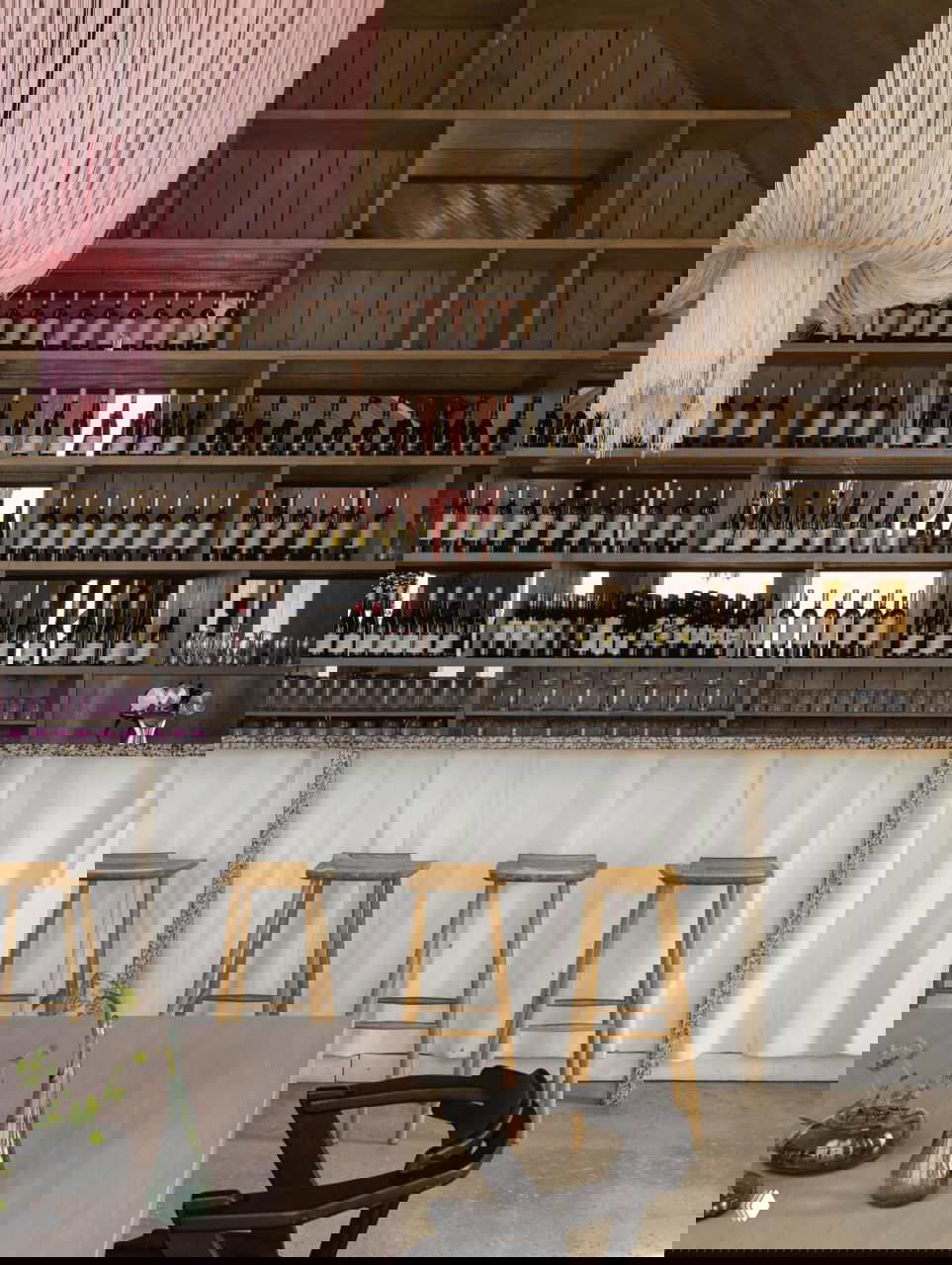 Барная зона ресторана с высокими табуретами: открытые деревянные полки, множество бутылок вина и крупная розовая текстильная люстра над стойкой.