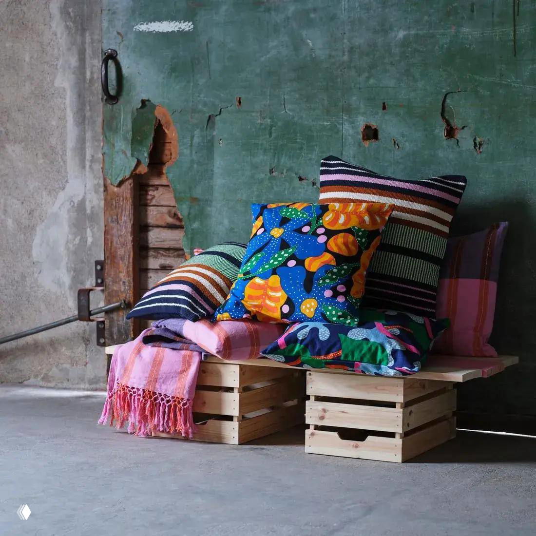 Стопка разноцветных подушек с яркими растительными и полосатыми принтами, покрывало и деревянные ящики на грубой стене — игра фактур и цвета.
