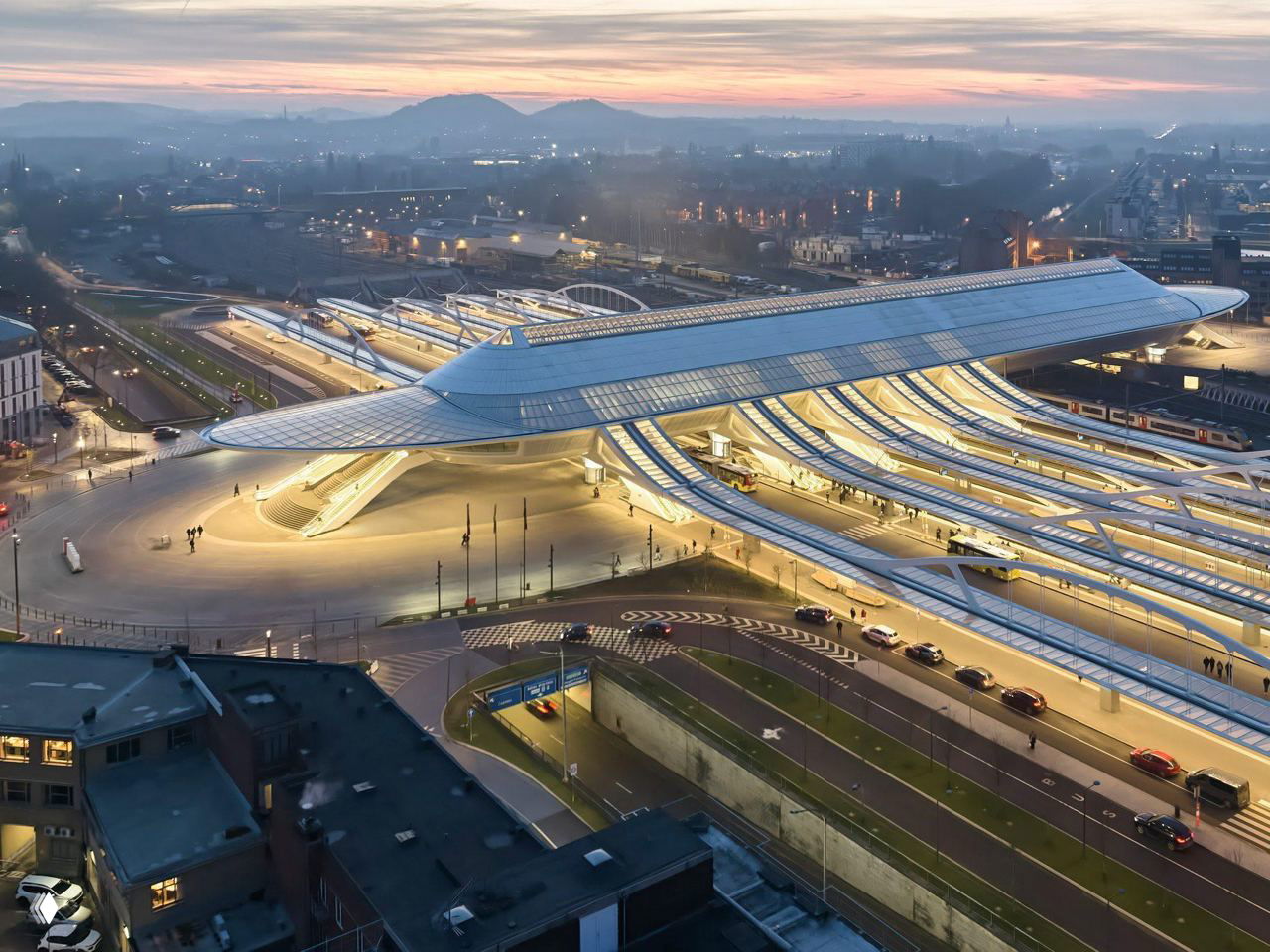 Панорамный вид на новый железнодорожный вокзал-мост в Монсе: длинная стеклянно-стальная волнообразная крыша, освещённая вечером над подъездными путями.