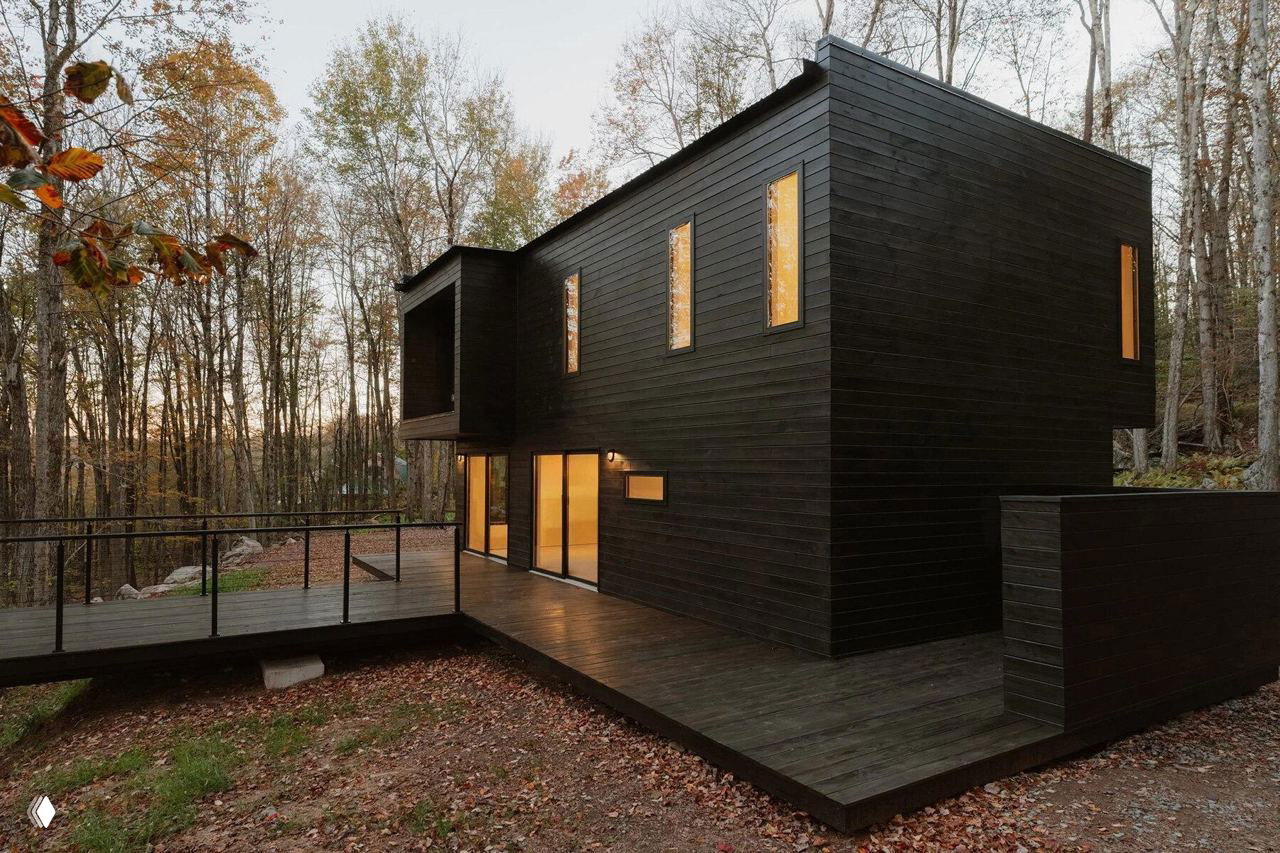 Угол дома и терраса: тёмный облицовочный фасад, большие панорамные окна с тёплым вечерним освещением, деревянный настил террасы и опавшая листва вокруг на лесистом участке.