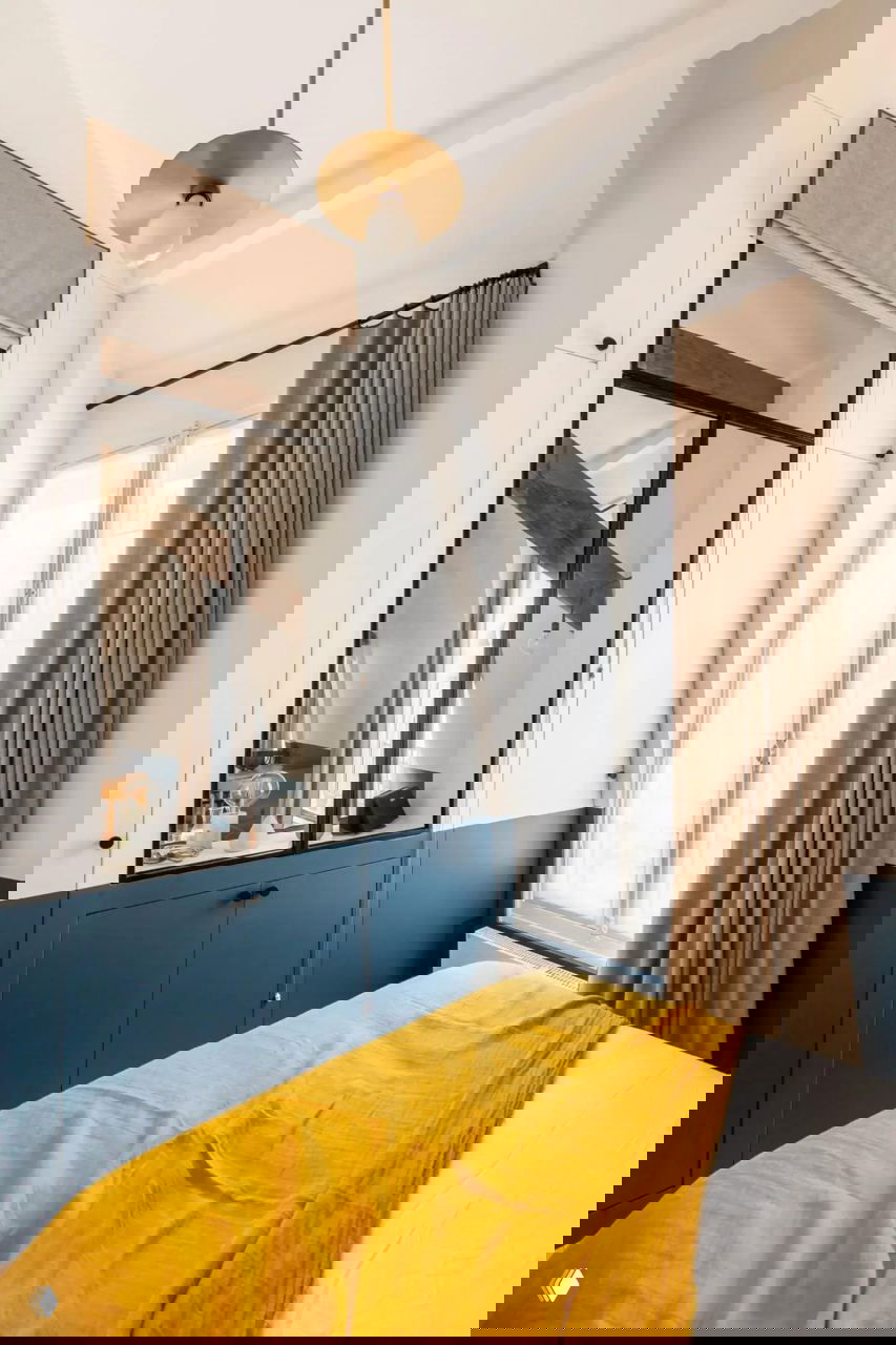 Спальня и окно с зеркальной стеклянной перегородкой, бежевые шторы, синяя нижняя покраска и солнечное жёлтое покрывало на кровати в компактном парижском интерьере.