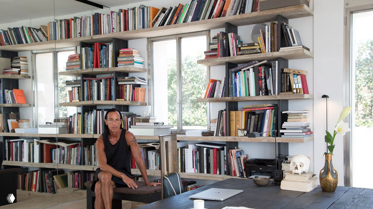 Мужчина сидит в светлой домашней студии рядом с открытыми деревянными полками, множеством книг и рабочим столом у окна.