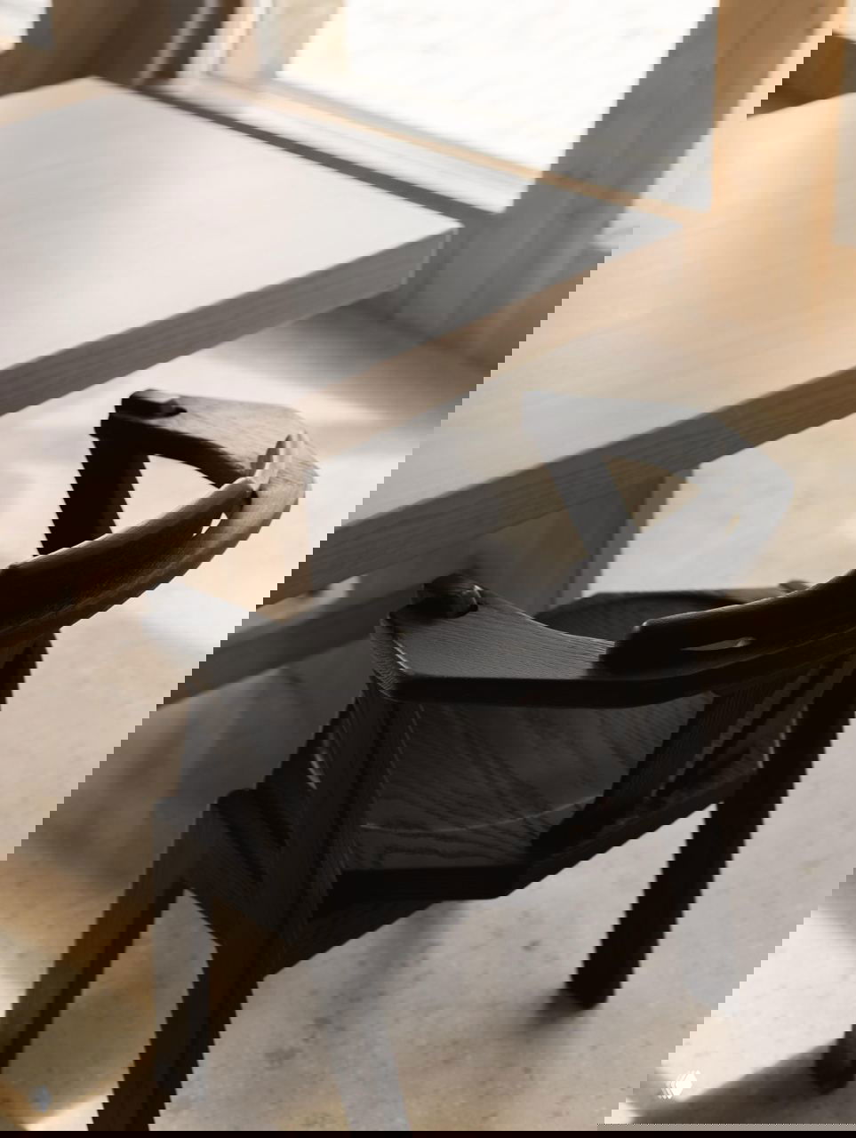 Крупный план: чёрный деревянный стул и светлый деревянный стол у окна, выраженная фактура материалов и сдержанная цветовая гамма интерьера ресторана.