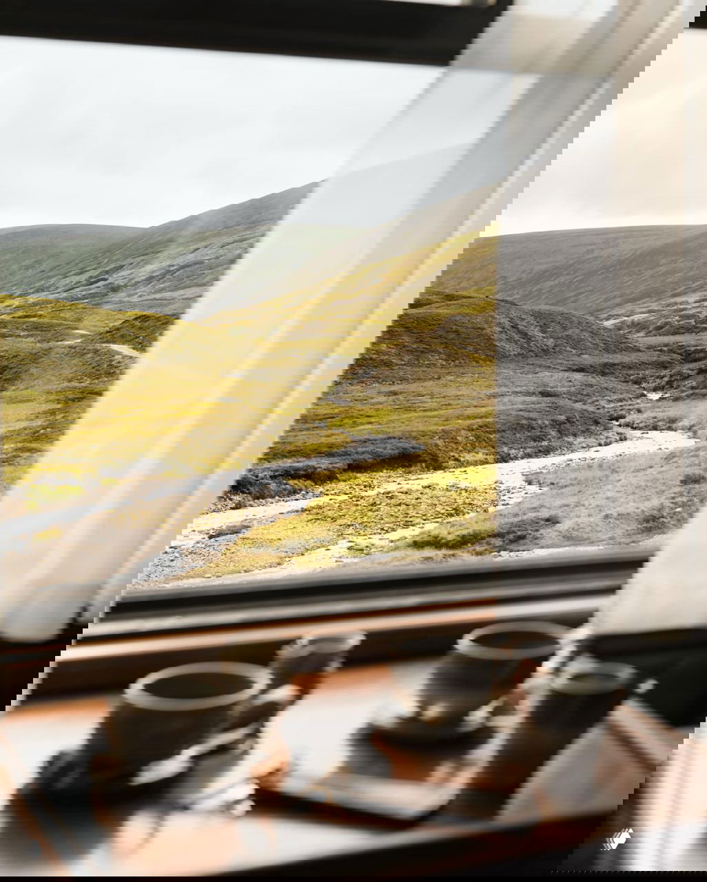 Вид из купе на шотландские холмы и ручей через окно; на подоконнике чашки и сервировка, создающие камерную атмосферу поездки по природе