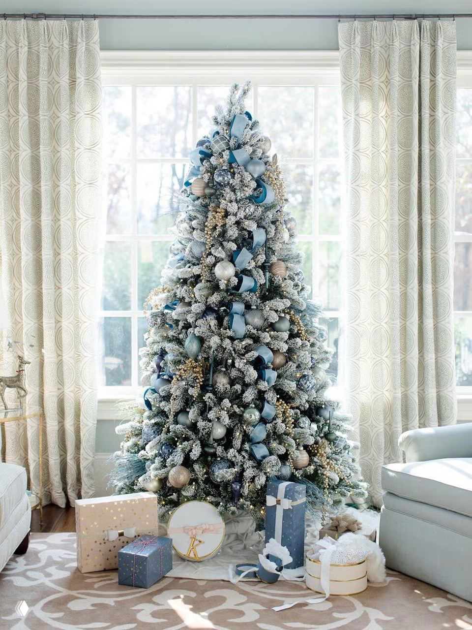 Снежно‑покрытая елка у большого окна, украшенная серебристо‑голубыми лентами, шарами и подсветкой, рядом подарки и светлый уютный интерьер.