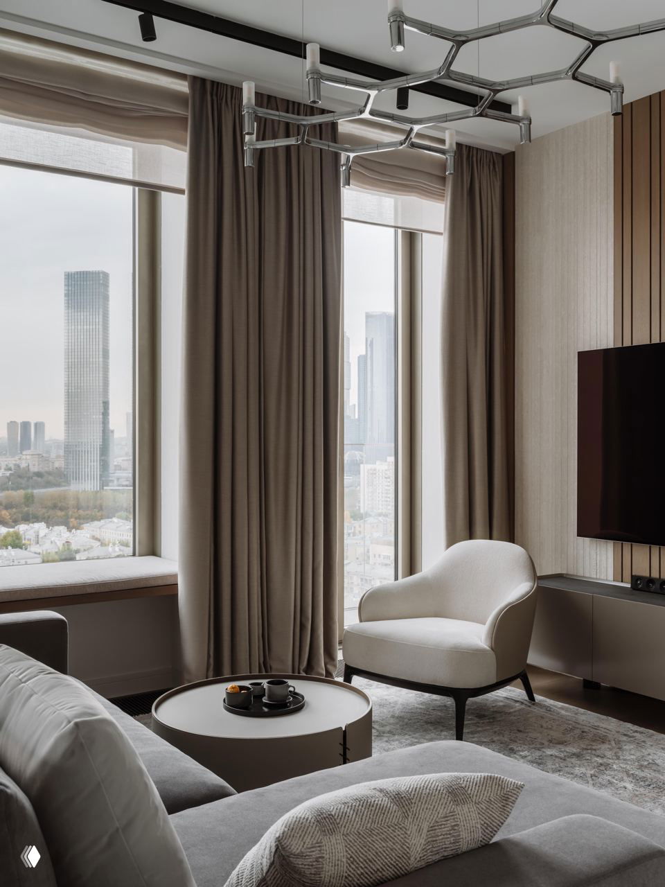 Уютный угол в гостиной: мягкое кресло у окна, низкий столик и длинные шторы в спокойных бежево‑серых тонах, характерных для джапанди‑эстетики.