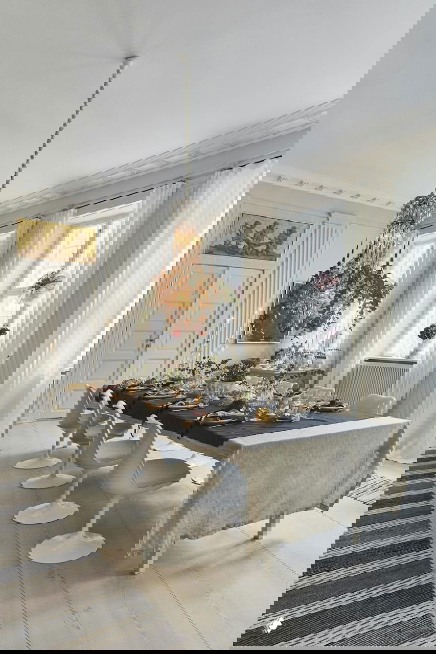 Общий вид столовой в квартире в Копенгагене: длинный сервированный стол, барочная люстра, высокие окна и светлые шторы.