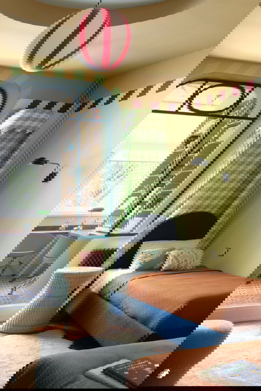 Одна из кроватей с зелёным клетчатым балдахином, оранжевым покрывалом и плетёным пуфом рядом; на заднем плане большой рабочий стол у окна.