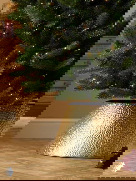 Крупный план основания рождественской ёлки: золотая металлическая чаша-основание на деревянном полу, тёплое освещение и иголки ветвей.