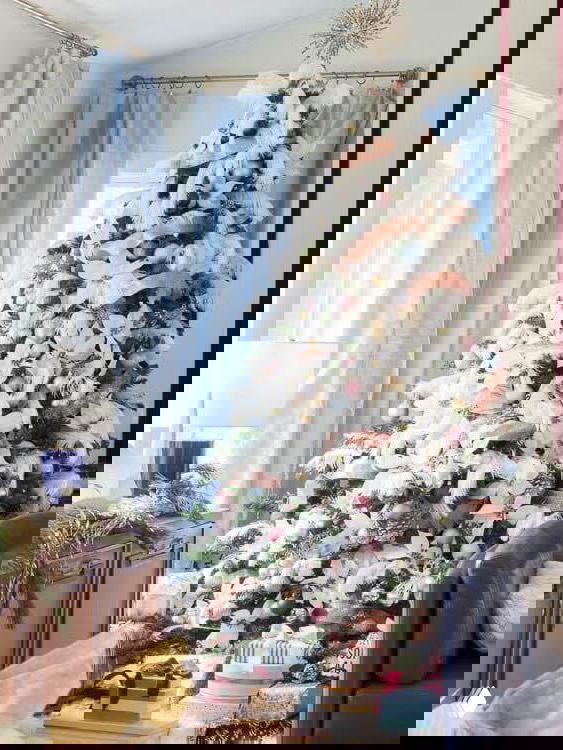 Тонкая высокая елка в светлом помещении, украшенная пастельными помпонами и подставкой с подарками, создающая мягкий и игривый праздничный образ интерьера.