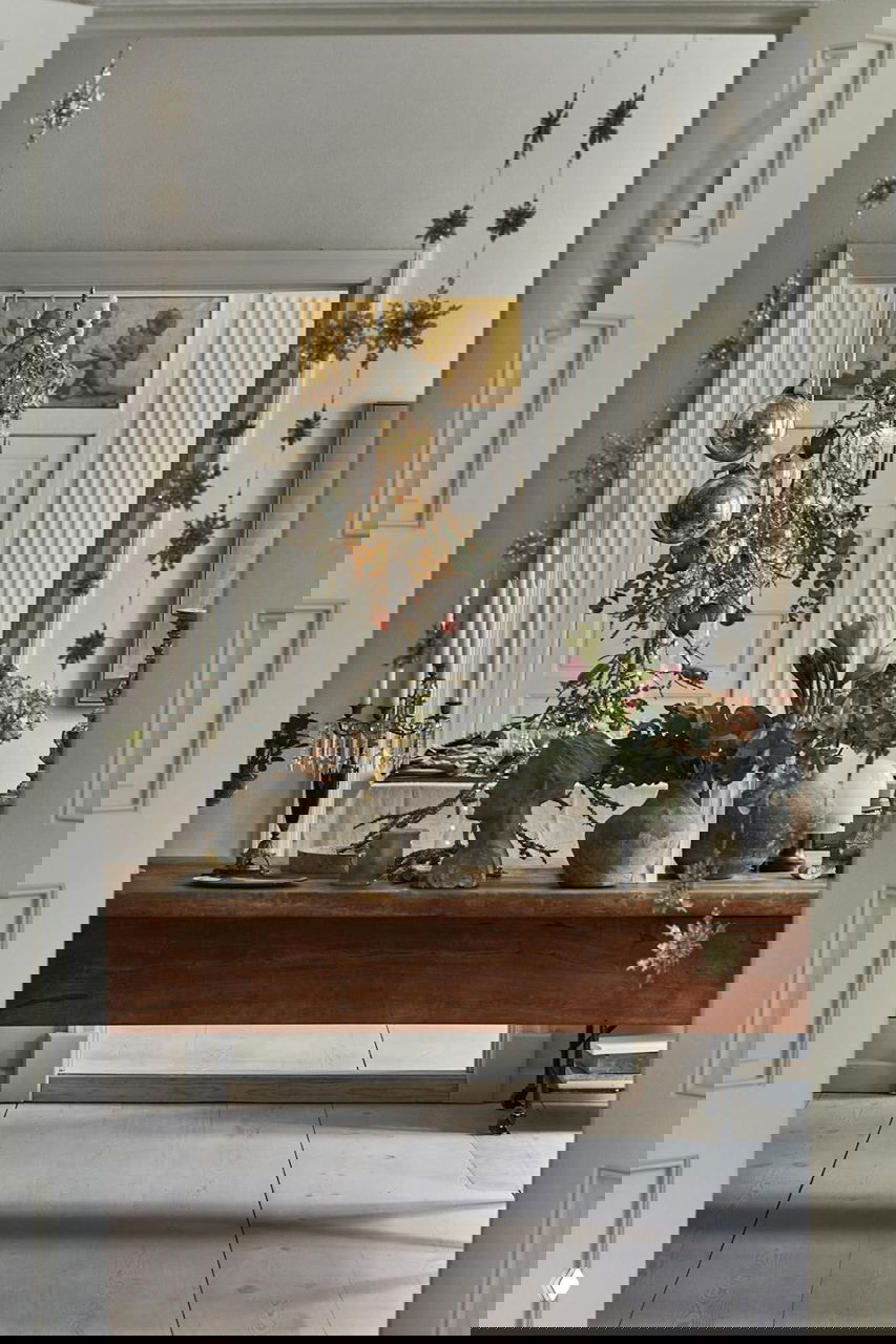 Вид через дверной проем на сервированный стол и подвешенную люстру с елочными шарами; крупные вазы и скульптурные детали на консоли.