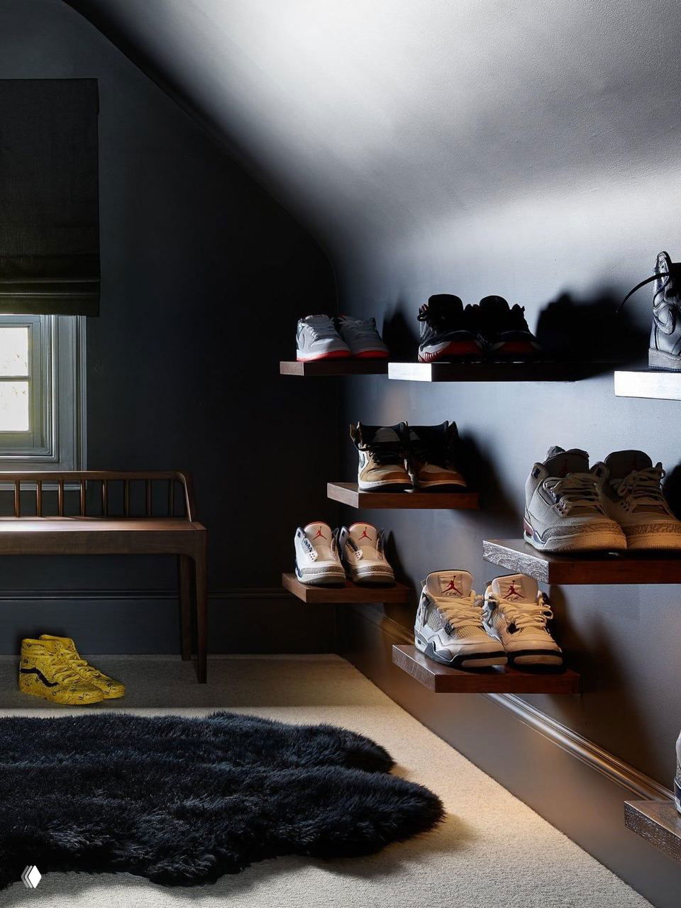 Настенные плавающие полки из тёмного дерева, на которых расставлены кроссовки и ботинки в контрастном минималистичном интерьере с мягким освещением.