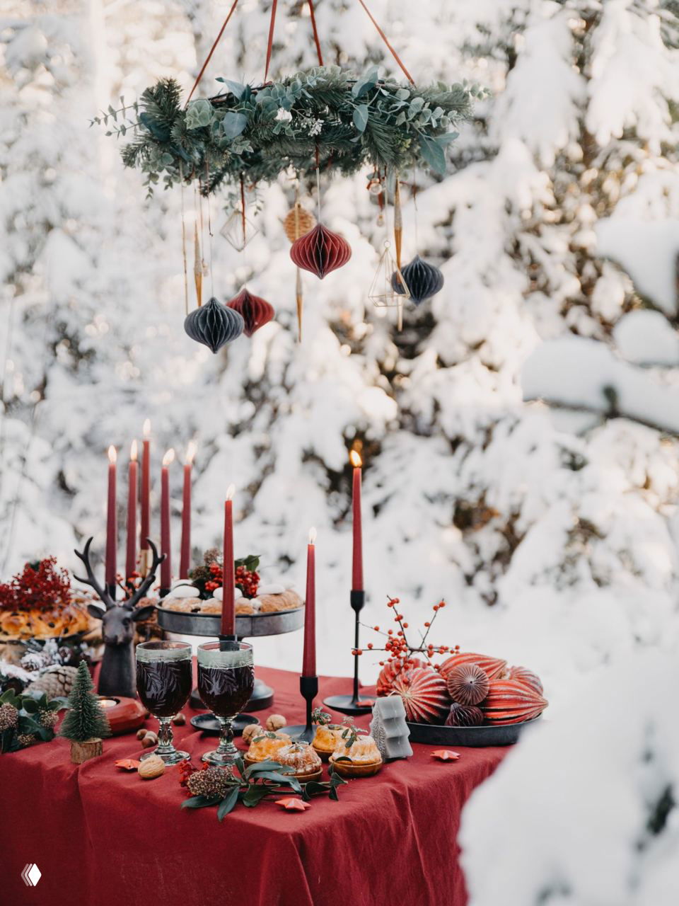 Уличный зимний стол под рождественским венком: красные свечи, бумажные украшения и праздничный натюрморт на фоне снега