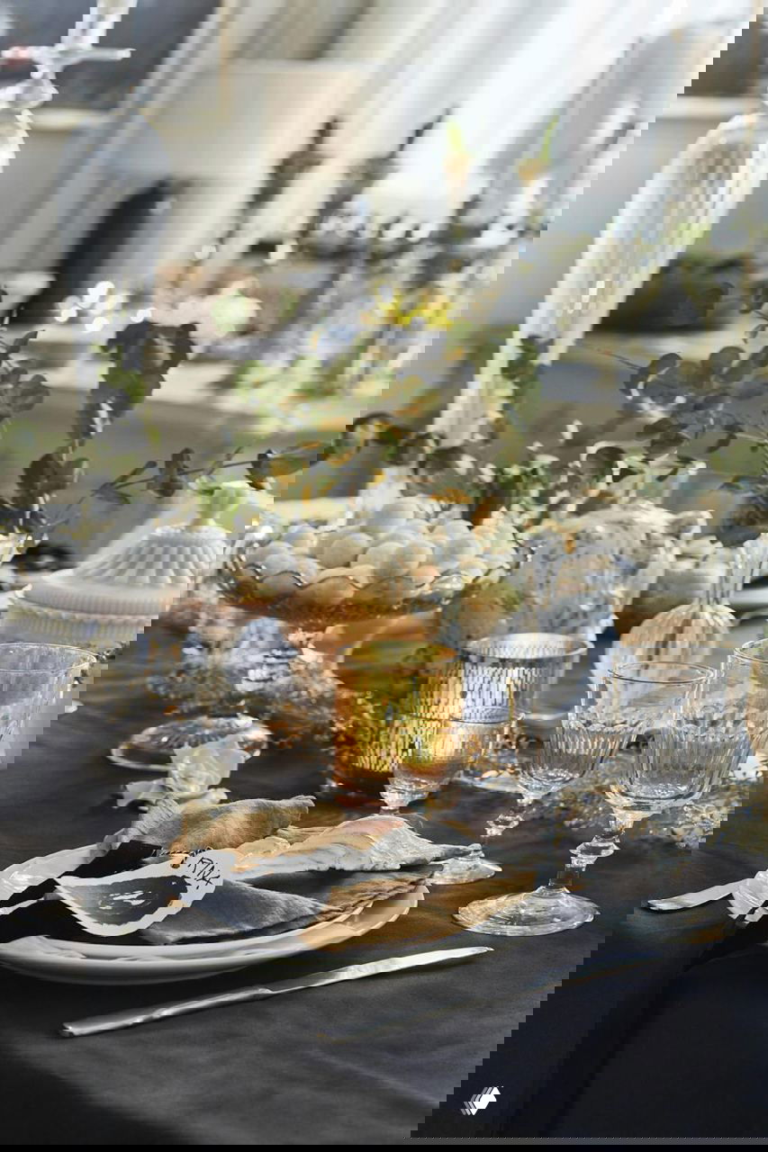 Крупный план праздничного стола: белый фарфор, хрустальные бокалы, льняные салфетки и свечи на темной фактурной скатерти.