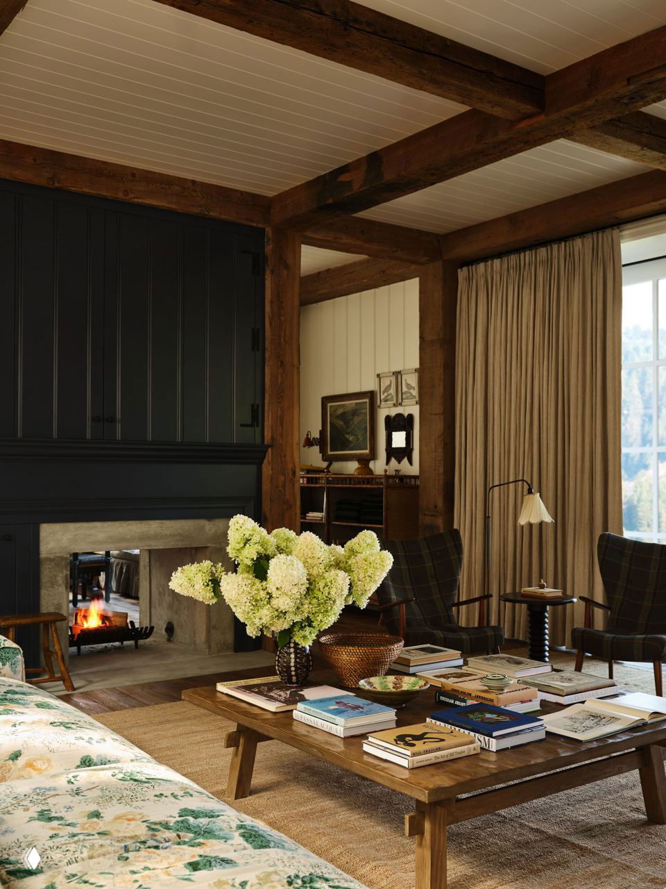 Просторная гостиная с деревянными балками, камином и большим журнальным столом, усыпанным книгами и вазой с белыми цветами, тёплая палитра тканей.