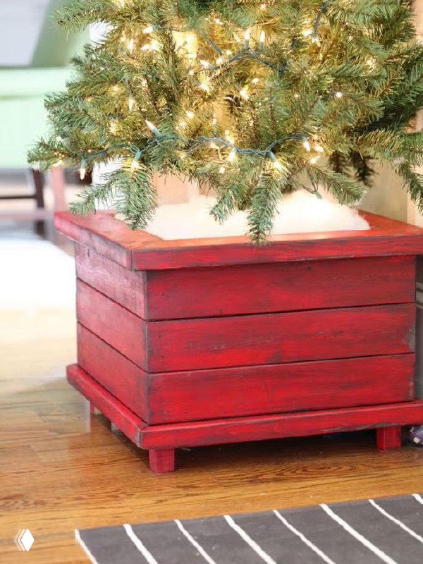 Красный деревянный ящик как подставка под ёлку: крупный план покрашенной древесины, ножки и топ с искусственным снегом вокруг ствола.