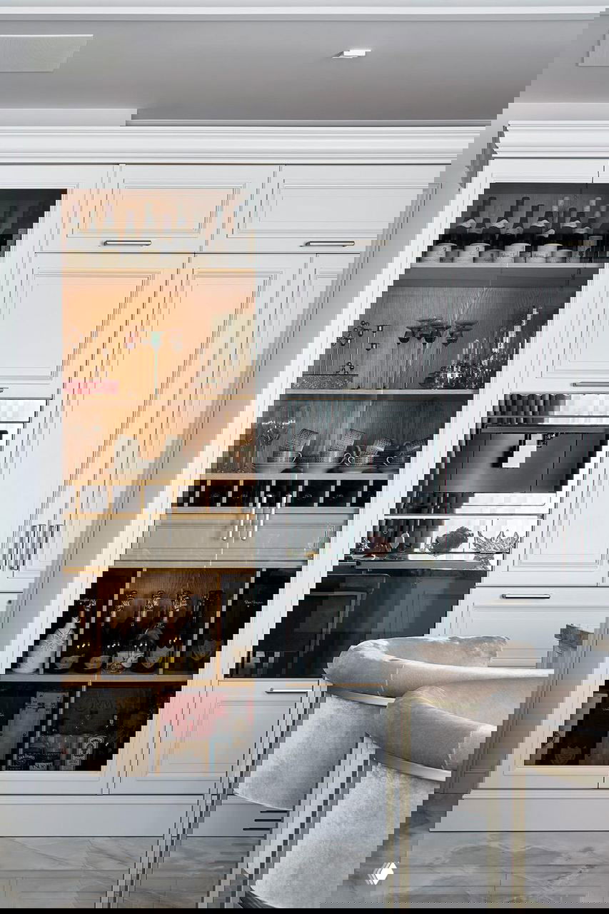 Кухня-столовая: белые классические фасады, встроенная техника и стеклянная витрина с коллекцией винных бутылок и подсветкой внутри шкафа.