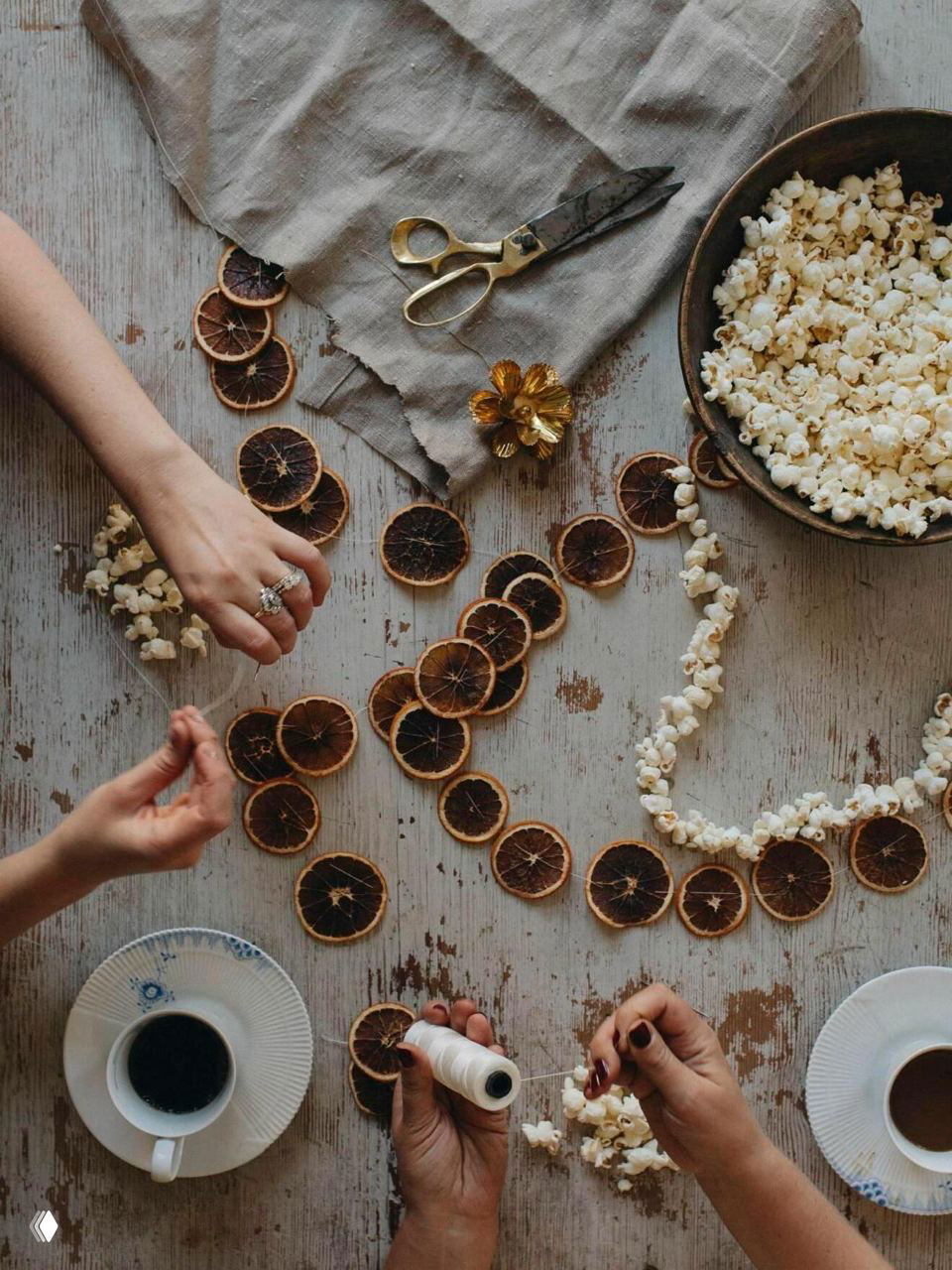 Плоская композиция: люди нанизывают попкорн и сушёные кружочки апельсина на нить для изготовления съедобной гирлянды на деревянном столе с чашками кофе.