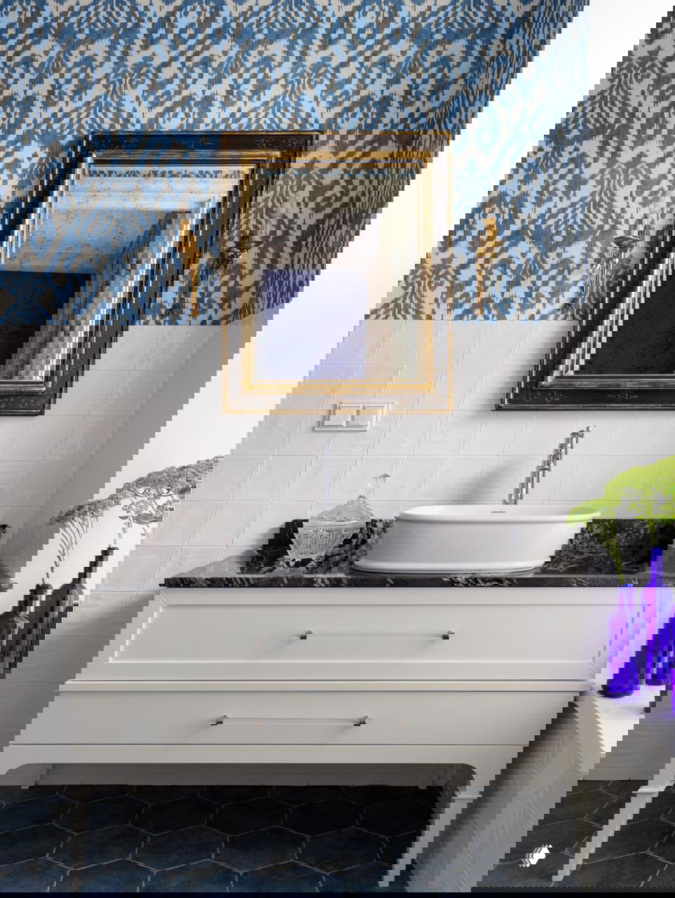 Раковина и белая мебельная тумба на фоне плитки в стиле икат и синих шестигранных полов, зеркало в золочёной раме и сдержанные декоративные акценты.