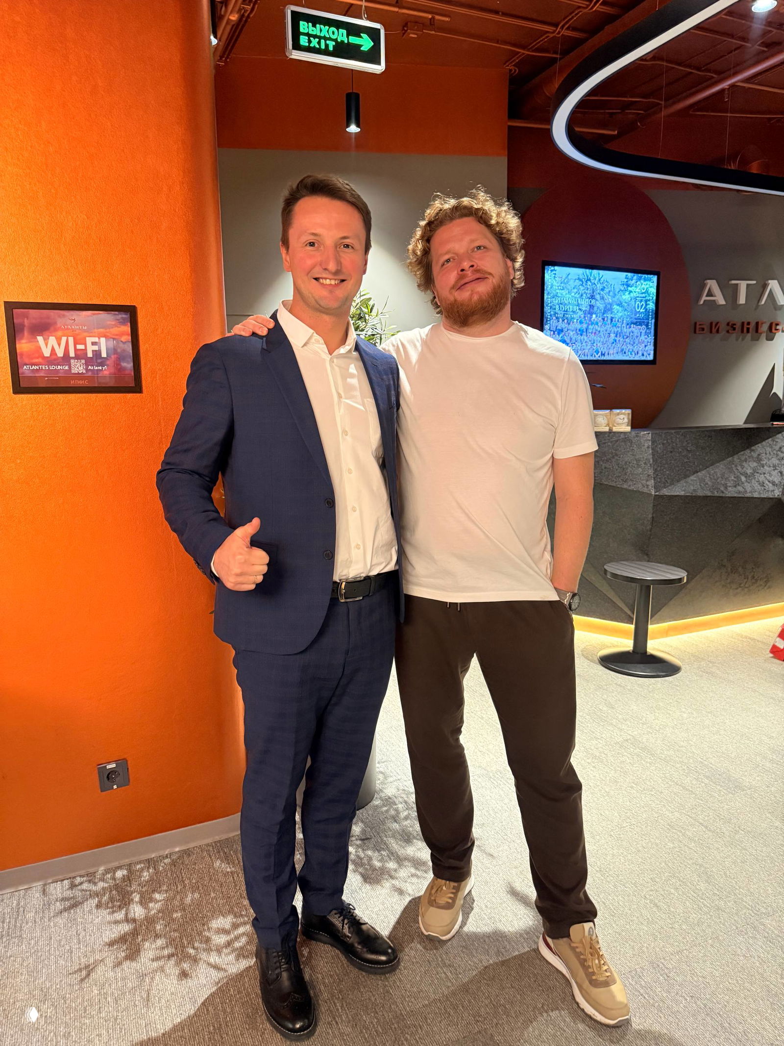 Двое мужчин в фойе бизнес‑клуба с оранжевой стеной; один в деловом костюме, второй в повседневной одежде — фото после мастер‑класса в Атлантах.