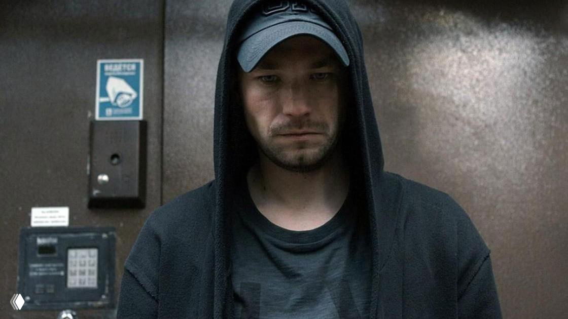 Мужчина в тёмной худи и кепке стоит в замкнутом пространстве с опущенным взглядом — образ задумчивости и изоляции, иллюстрация стигмы и сокрытия ВИЧ‑статуса.