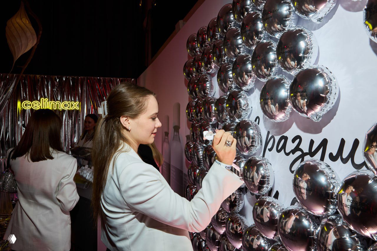 Гость подписывает серебристые шары на стенде; вокруг другие посетители и брендовая инсталляция с логотипом мероприятия.