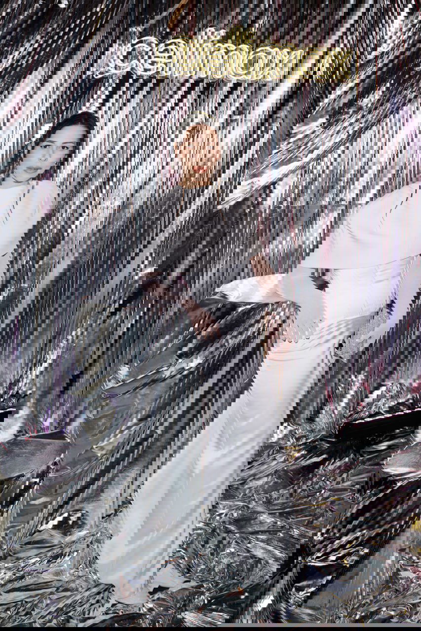 Женщина в белом платье позирует у серебристой фотозоны с логотипом партнёра; сцена оформлена глянцевыми тканями и декоративными элементами.