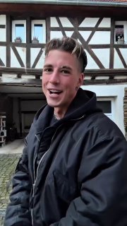 Молодой мужчина на улице у фахверкового дома улыбается в камеру — уличный кадр из ролика тренда о танцах под «Катюшу».