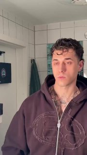 Крупный план мужчины в тёмной толстовке в помещении с зеркалом и крючками на стене; кадр из короткого видео в формате вертикального ролика.