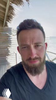 Портрет мужчины с короткой стрижкой и бородой на фоне пляжа и деревянной постройки; дневной портрет в естественном освещении.