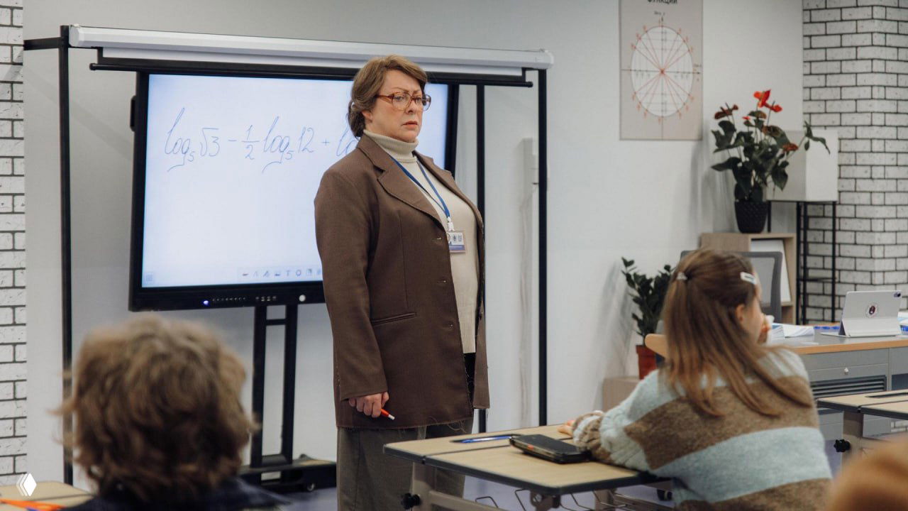 Мария Аронова в роли учительницы математики в классе перед интерактивной доской, кадр из комедийного сериала «Олдскул», учащиеся на переднем плане