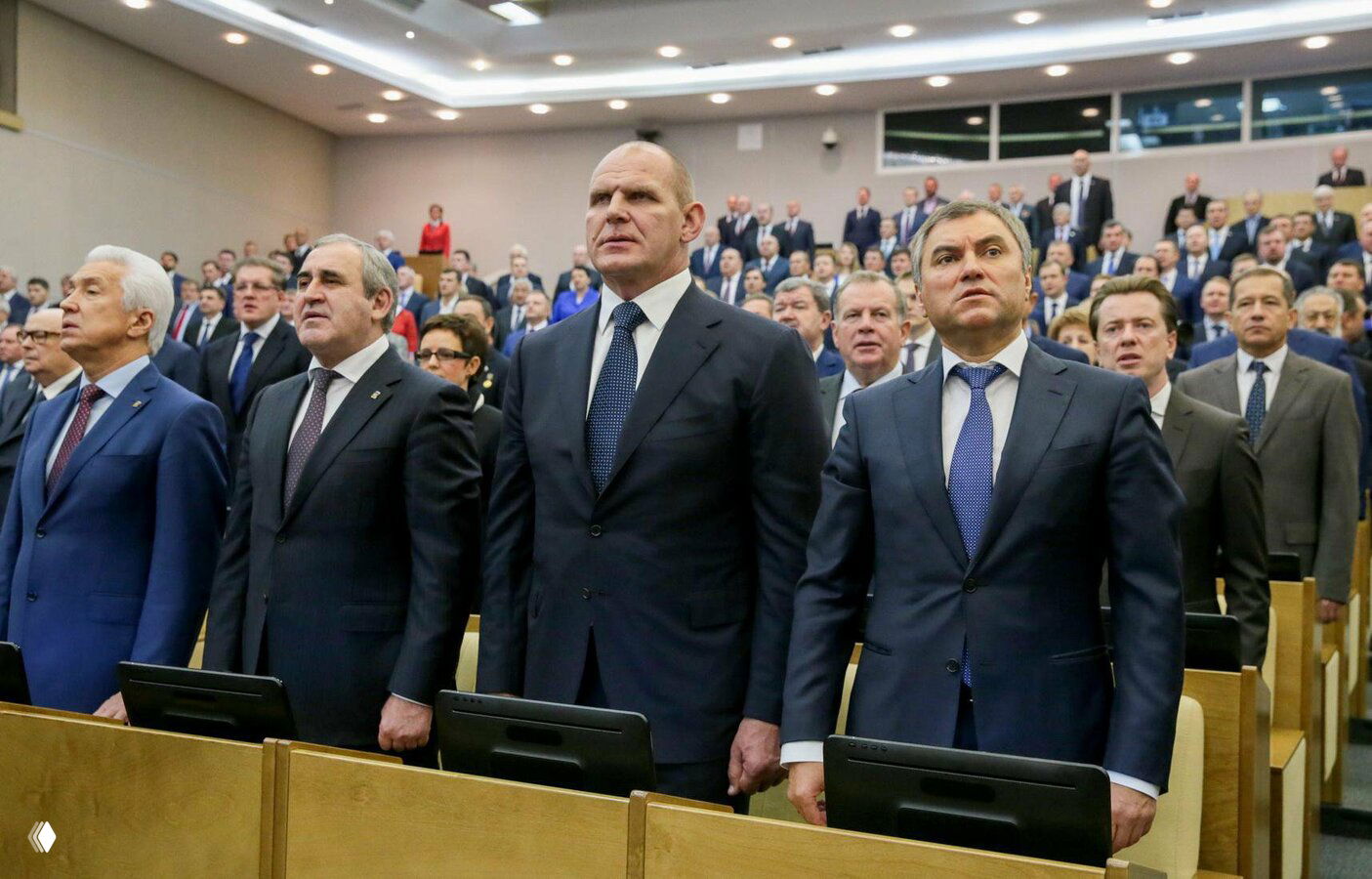 Группа депутатов в строгих костюмах и галстуках стоит в зале Государственной думы во время заседания; общий план, много участников.