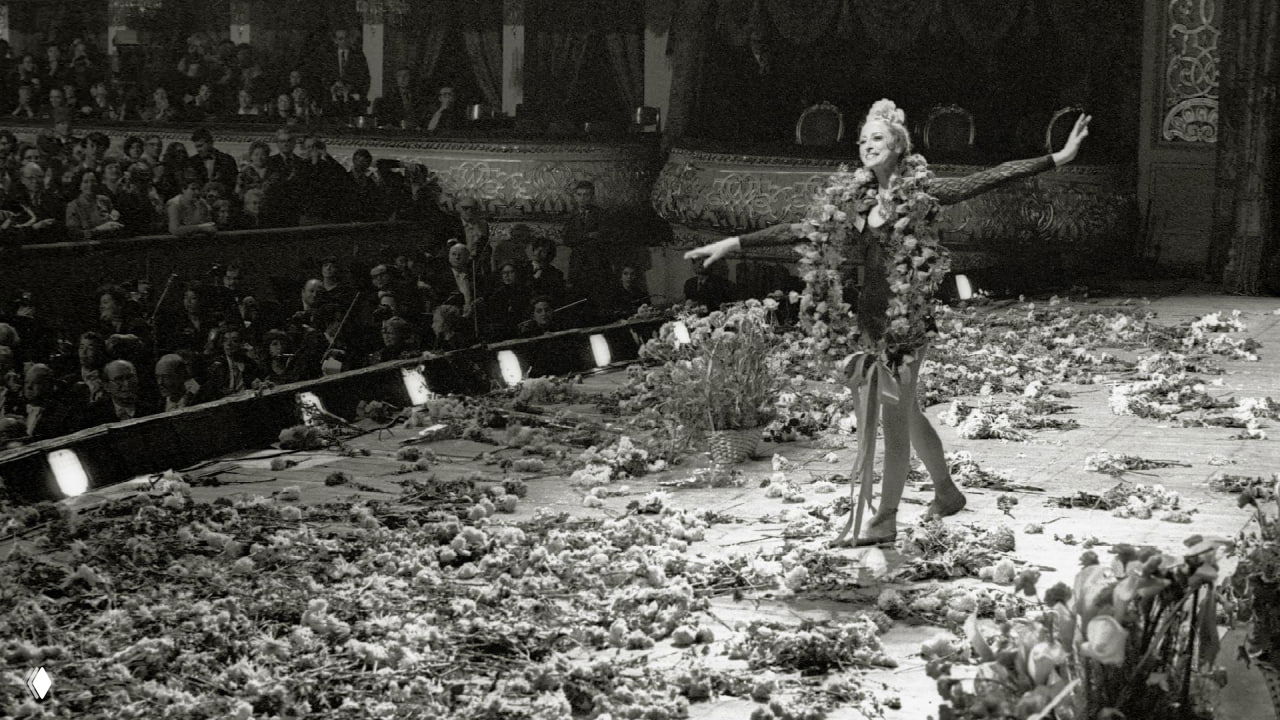 Чёрно‑белая фотография Майи Плисецкой на сцене во время поклона, сцена усыпана цветами и букетами, публика аплодирует