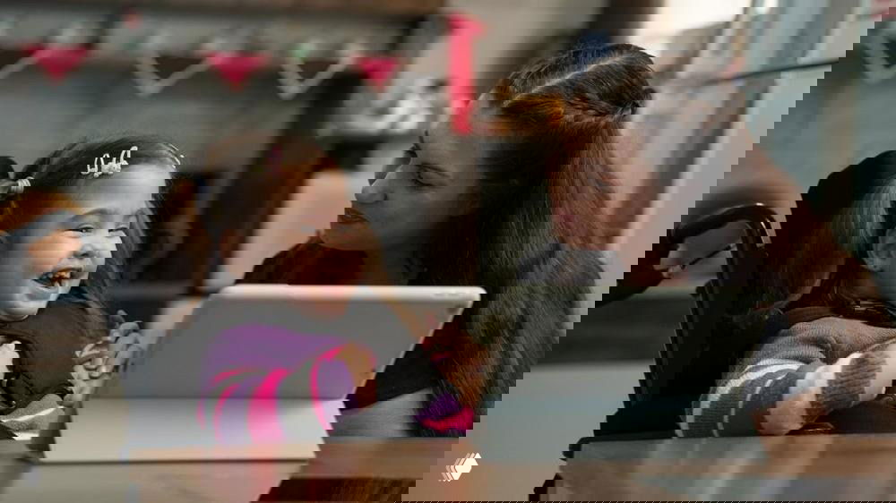 Мама смотрит на улыбающуюся девочку в инвалидной коляске за столом рядом с планшетом; тёплая домашняя сцена поддержки и заботы.