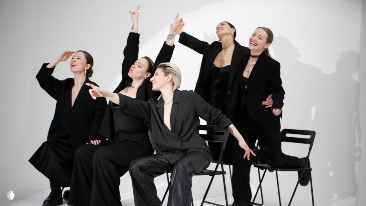 Кадр: актрисы в чёрных костюмах сидят и стоят на стульях в студии — сцена спектакля о принятии тела.