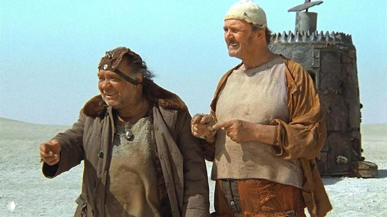 Кадр из фильма «Кин‑дза‑дза!»: двое персонажей в поношенной одежде на пустынном фоне рядом со странным металлическим сооружением, сцена фильма.