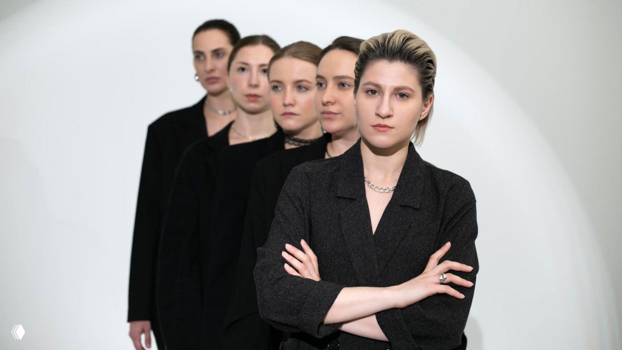 Групповая сцена: несколько актрис в строгих чёрных костюмах выстроены в линию; впереди актриса со скрещёнными руками