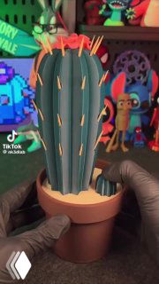 Фотография декоративного кактуса в горшке, выполненного как 3D-модель/печать, на рабочем столе с яркими фигурками и элементами декора на заднем плане.