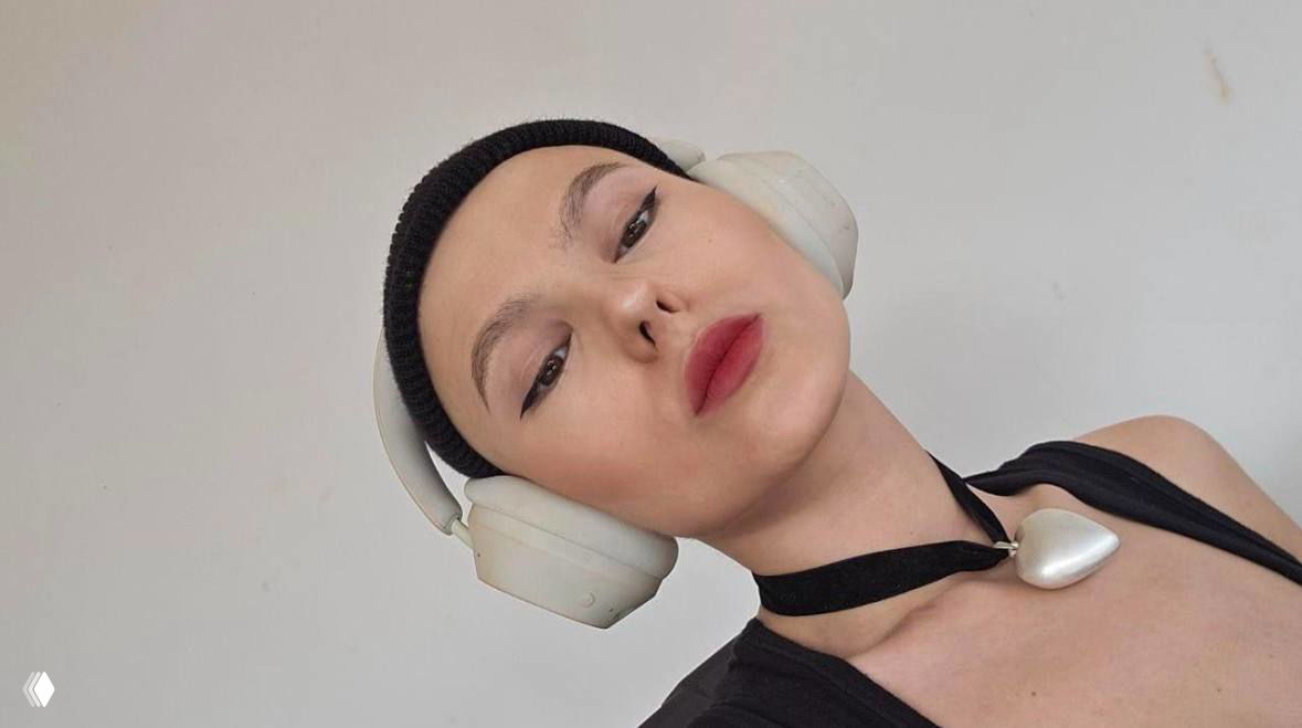 Портрет молодой женщины с короткой стрижкой и шапкой в белых наушниках; селфи из архива канала, лицо с макияжем и серьёзным выражением.