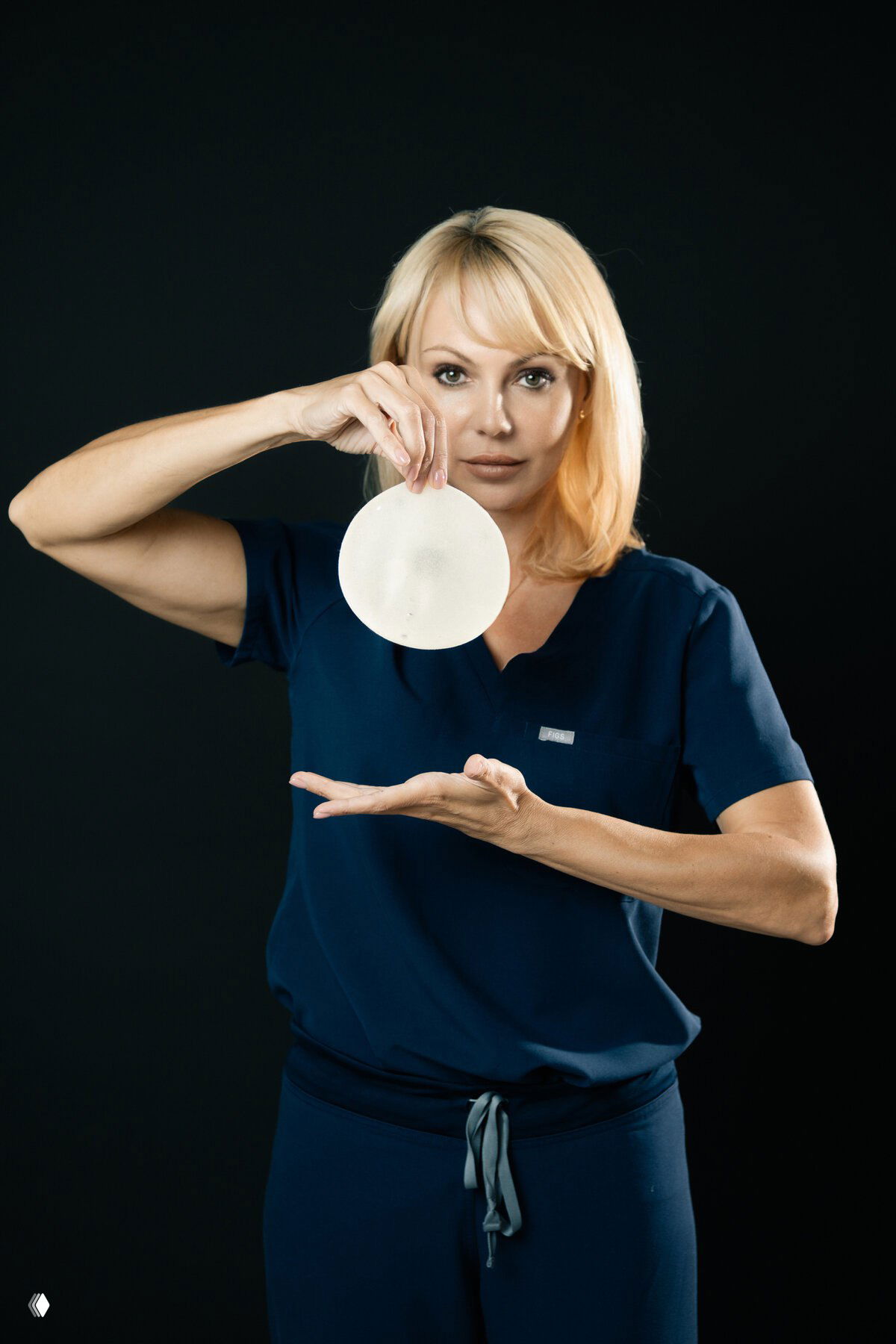 Пластический хирург в тёмно-синей форме демонстрирует круглый силиконовый грудной имплантат на тёмном фоне.