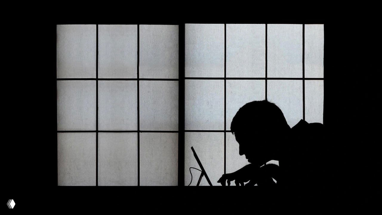 Чёрно-белый силуэт человека, наклонившегося над ноутбуком на фоне большого окна; образ изоляции, тревоги и таинственности.