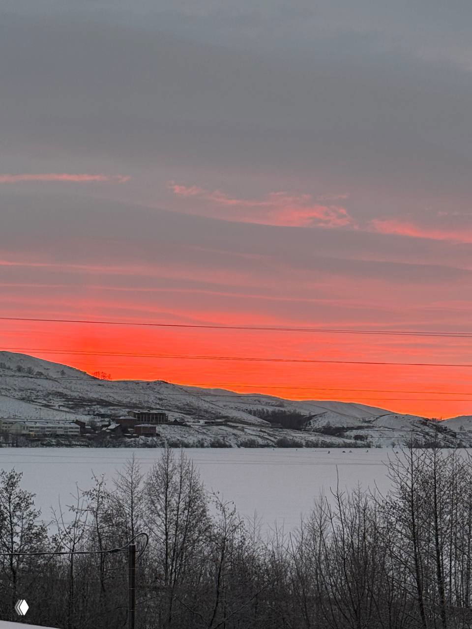 Яркий красный закат над замёрзшим озером и холмами, силуэты деревьев на переднем плане — атмосферный зимний пейзаж.