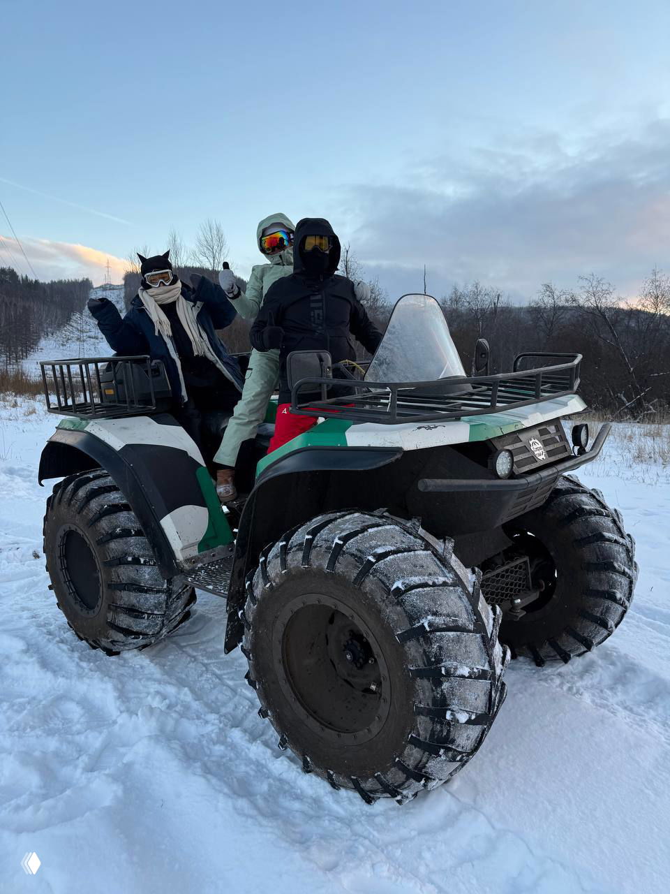 Крупный план квадроцикла с массивными шинами в снегу, на заднем плане участники в зимней экипировке готовятся к поездке.