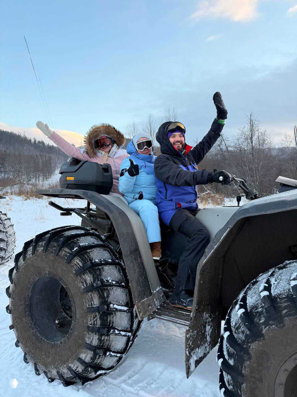 Трое участников в тёплой одежде радуются и машут руками, сидя на квадроцикле на фоне заснеженного ландшафта турбазы.