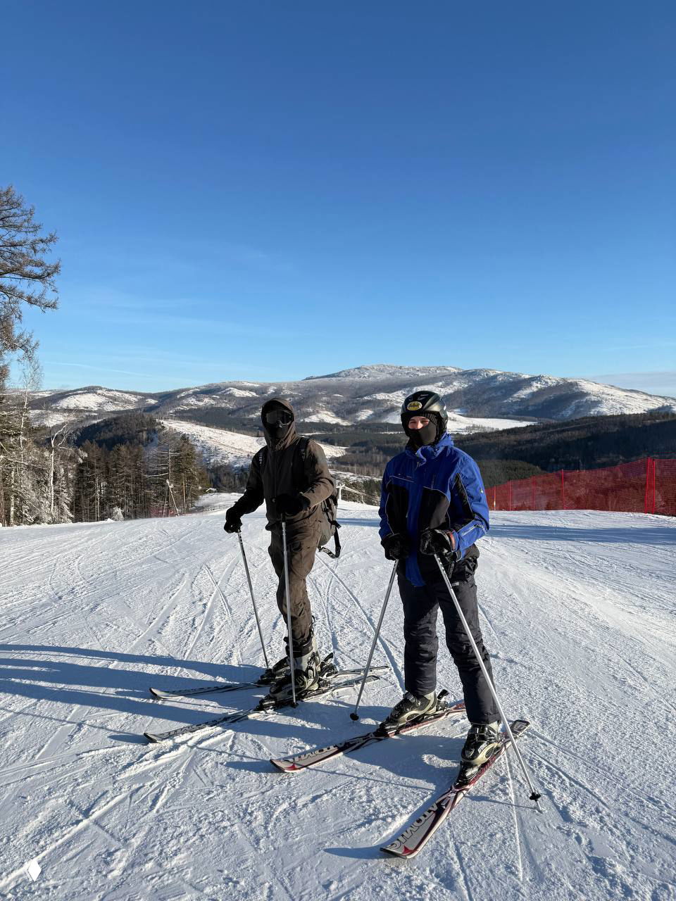 Два лыжника в масках и экипировке стоят на подготовленной трассе — вид на склон и дальние горы в солнечный зимний день.