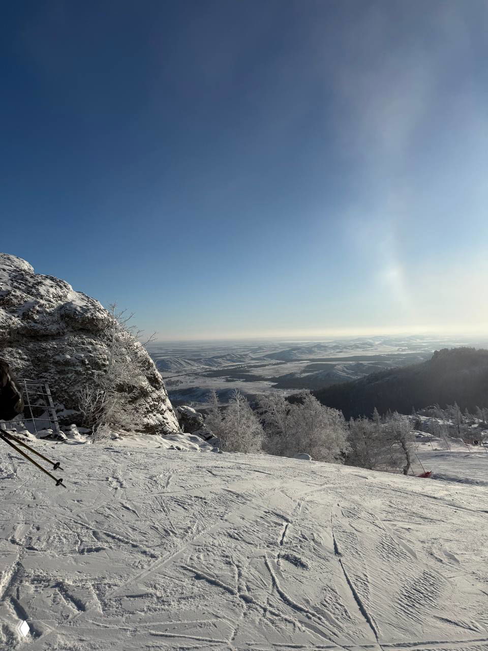 Заснеженный склон с отпечатками лыж и открытой долиной внизу, спокойное голубое небо и перспективный вид на заснеженные холмы.