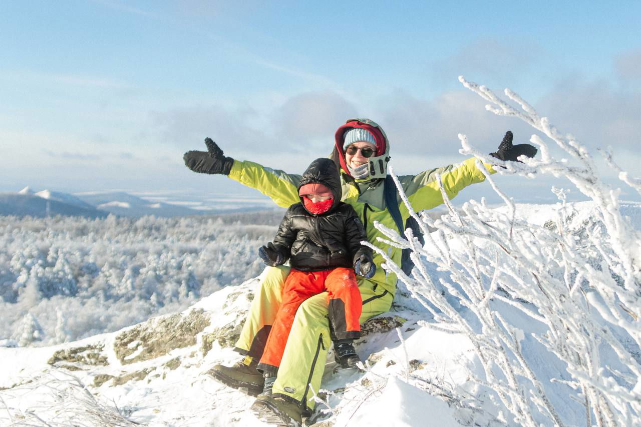 Двое туристов на заснеженной скале: взрослый и ребёнок в яркой одежде улыбаются на фоне облеплённых инеем ветвей и далёких вершин.