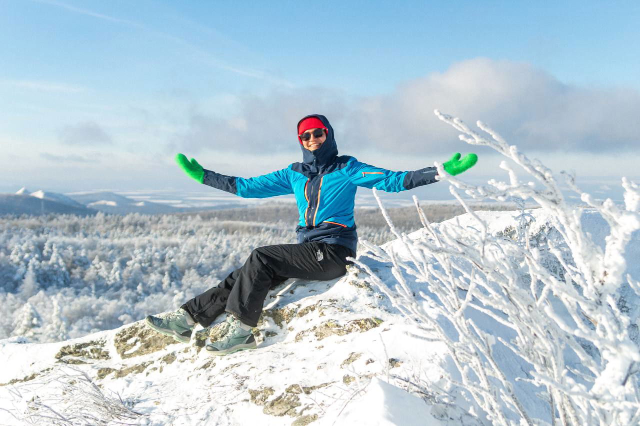 Турист в синей куртке и зелёных варежках сидит на заснеженной скале и обнимает воздух, вокруг белый лес и ясное зимнее небо над горами.