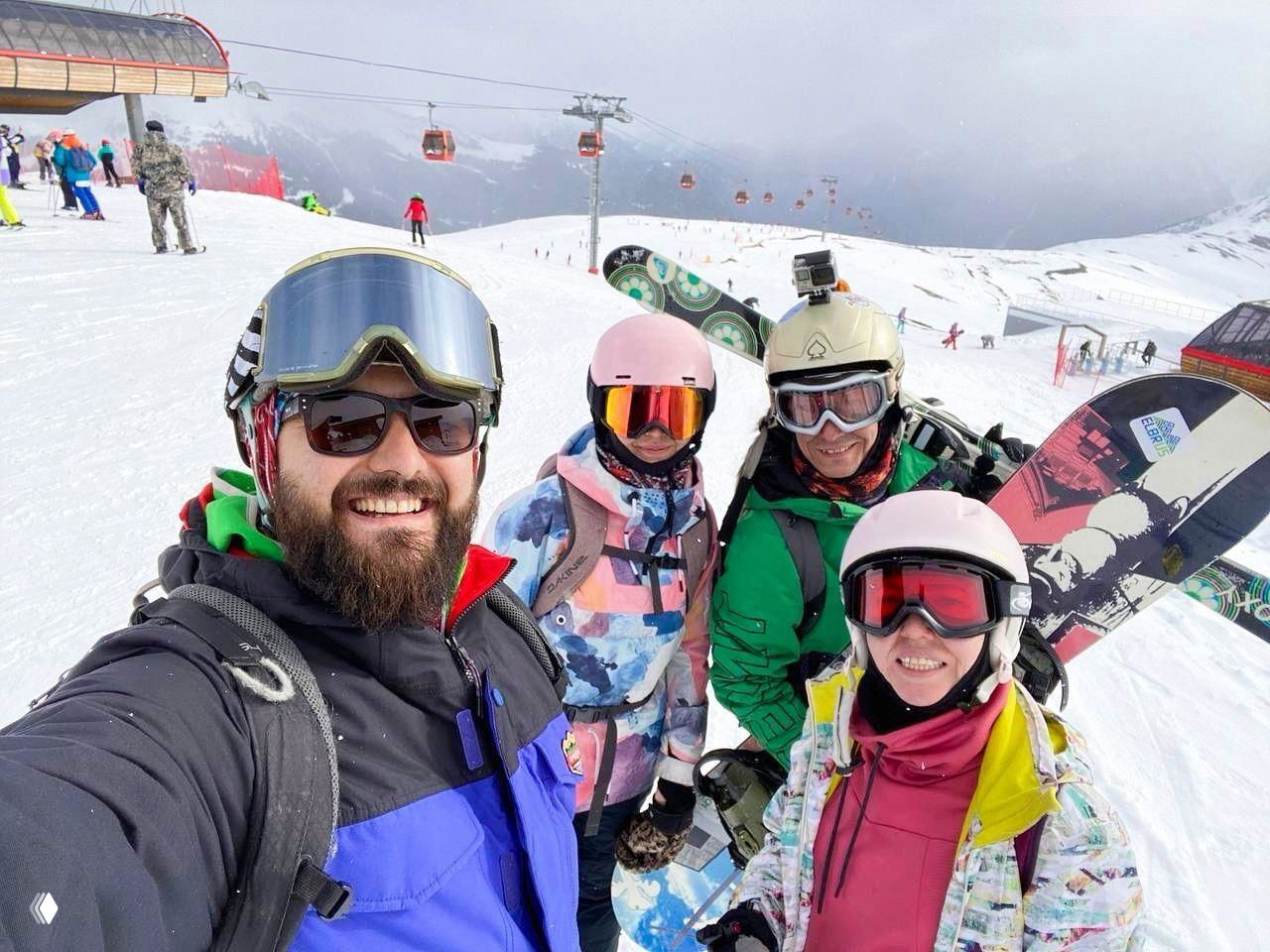 Группа лыжников и сноубордистов делает селфи на снежном склоне; на заднем плане видны подъёмники и горные вершины, улыбки участников тура.