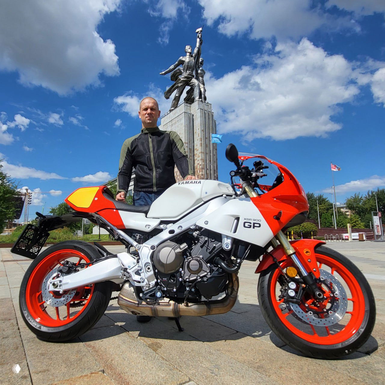 Yamaha XSR900 GP в боковом ракурсе на городской площади: оранжево‑белый мотоцикл с яркими колёсами, рядом стоит автор на фоне памятника и синего неба.